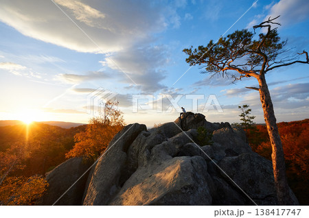 Sun sets, casting golden glow over rocky landscape and autumnal trees. Lone person atop rocks, silhouetted against vibrant sky. Dovbush Rocks, Carpathian mountains, Ukraine. 138417747