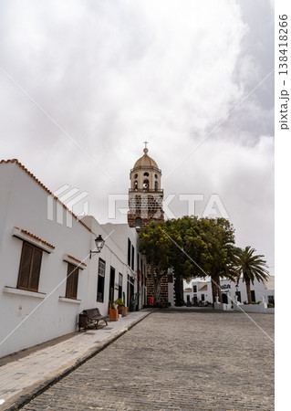 A view of the historic church bell tower in Teguise, Lanzarote, framed by the white buildings and the cobblestone street. A beautiful travel destination. A view of the historic church bell tower in Teguise, Lanzarote, framed by the white buildings and the cobblestone street. A beautiful travel destination. 138418266