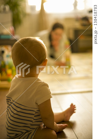 Caucasian Baby Sitting On Floor Watching Blurred Caregiver In Background, Warm Backlit Scene, Reflective Pause, Domestic Playroom Mood And Family Connection verical Caucasian Baby Sitting On Floor Watching Blurred Caregiver In Background, Warm Backlit Scene, Reflective Pause, Domestic Playroom Mood And Family Connection verical 138418783