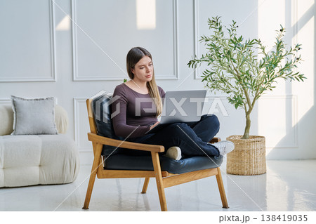 Portrait of young woman sitting on chair with laptop, in home interior 138419035