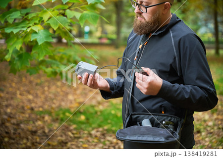 A man with a beard inspects a small device in an autumn park. Colorful leaves cover the ground, and he is focused on the gadget as he balances a bag on his side. A man with a beard inspects a small device in an autumn park. Colorful leaves cover the ground, and he is focused on the gadget as he balances a bag on his side. 138419281