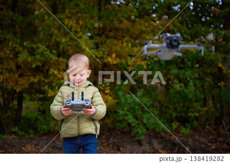 A young child stands in a lush park surrounded by vibrant autumn leaves, skillfully maneuvering a drone with a remote control. Excitement fills the air. A young child stands in a lush park surrounded by vibrant autumn leaves, skillfully maneuvering a drone with a remote control. Excitement fills the air. 138419282