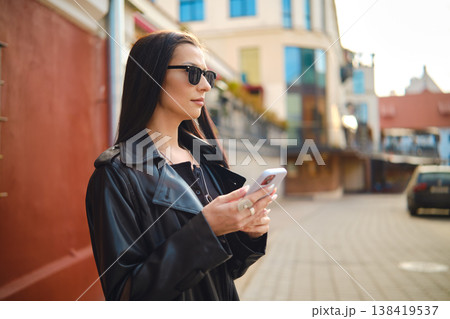 Young woman checks her phone while standing on city street in afternoon sunlight Young woman checks her phone while standing on city street in afternoon sunlight 138419537