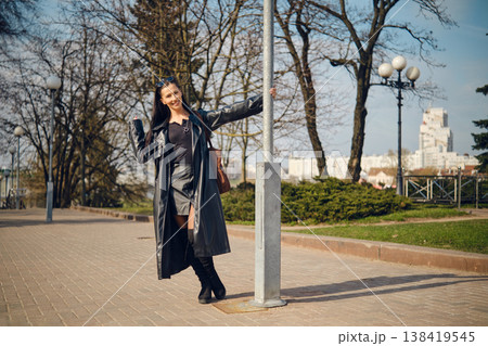 Woman stands by a street lamp in a park during a sunny day in a city, smiling and waving at the camera Woman stands by a street lamp in a park during a sunny day in a city, smiling and waving at the camera 138419545