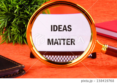 A gleaming golden magnifying glass reveals the phrase ideas matter on a bright orange surface. A verdant plant and a deep red book complete the creative scene 138419768