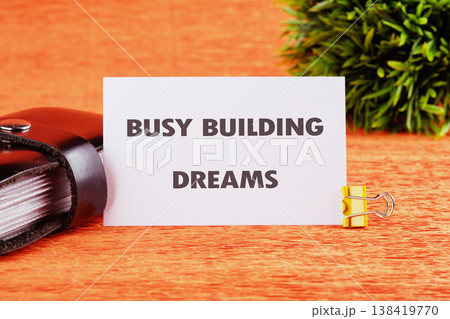 A crisp white card presents BUSY BUILDING DREAMS held by a yellow clip on a warm orange desk, alongside a dark notebook and lively green plant 138419770