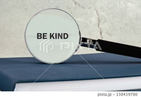 Magnifying glass on a dark blue book highlights BE KIND on a grey surface in daylight Magnifying glass on a dark blue book highlights BE KIND on a grey surface in daylight 138419780