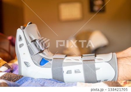 Foot in a medical boot rests on a bed with blankets during recovery at home after an injury 138421705