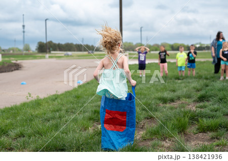 Child runs in sack race during outdoor activity in a park on a cloudy day with other kids watching Child runs in sack race during outdoor activity in a park on a cloudy day with other kids watching 138421936