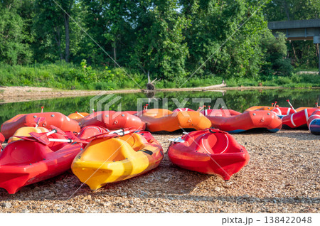 People prepare for kayaking on a warm sunny day at a river with red and yellow kayaks lined up on the shore 138422048