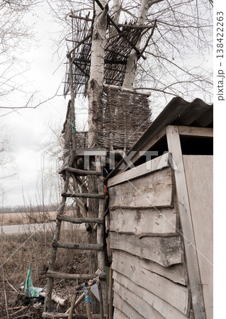 Rustic Treehouse Constructed from Natural Branches and Woven Twigs Beside Weathered Wooden Shed Under Overcast Sky with Dry Grass Field Background Rustic Treehouse Constructed from Natural Branches and Woven Twigs Beside Weathered Wooden Shed Under Overcast Sky with Dry Grass Field Background 138422263