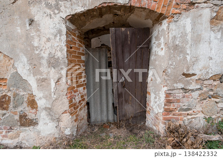 Weathered Brick Archway with Rustic Wooden Door in Abandoned Stone Building Showing Peeling Plaster and Corrugated Metal Interior Detail Weathered Brick Archway with Rustic Wooden Door in Abandoned Stone Building Showing Peeling Plaster and Corrugated Metal Interior Detail 138422332
