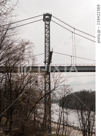 Tall Metal Suspension Bridge Tower Over River With Bare Trees And Grey Sky In Autumn Landscape Tall Metal Suspension Bridge Tower Over River With Bare Trees And Grey Sky In Autumn Landscape 138422369