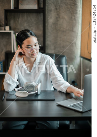 A young woman in a modern office setting, working on a laptop with headphones, showcasing a professional and focused work environment. 138424254