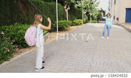 A mother and her school-age daughter wave farewell outside the school entrance. Back to school vibes A mother and her school-age daughter wave farewell outside the school entrance. Back to school vibes 138424832