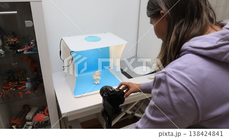 Person takes a picture of a small figure in a light box with a blue background inside a room filled with toys and small items in the background Person takes a picture of a small figure in a light box with a blue background inside a room filled with toys and small items in the background 138424841