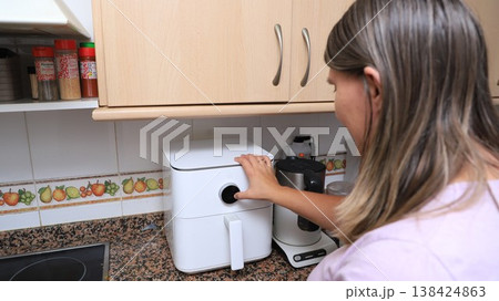 Woman operates an air fryer in a modern kitchen with wooden cabinets Woman operates an air fryer in a modern kitchen with wooden cabinets 138424863