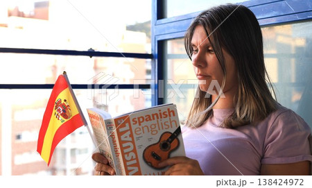 Young woman reading a Spanish-English visual dictionary while holding a small Spanish flag Young woman reading a Spanish-English visual dictionary while holding a small Spanish flag 138424972