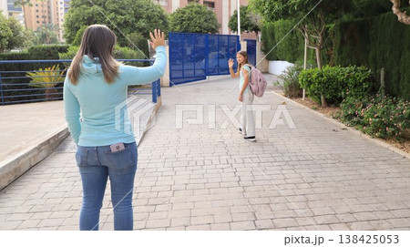 Mother waves goodbye to daughter at school gate, both smiling and ready for the day. Mother waves goodbye to daughter at school gate, both smiling and ready for the day. 138425053