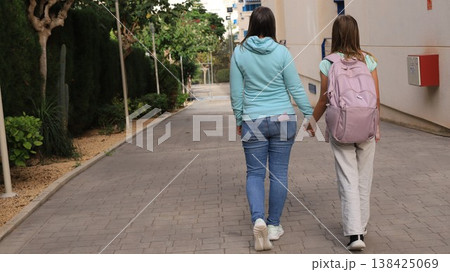 Caring mother in light blue hoodie holds hands with her young daughter carrying pink backpack 138425069