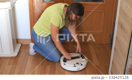 Woman repairs a robotic vacuum cleaner at home while kneeling on wooden floor near a door in daylight Woman repairs a robotic vacuum cleaner at home while kneeling on wooden floor near a door in daylight 138425126