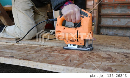 A jigsaw in the hand of a worker kneeling on a thick sheet of OSB and cutting the dense material into pieces, the sharp blade of the jigsaw cuts the massive OSB sheet 138426248