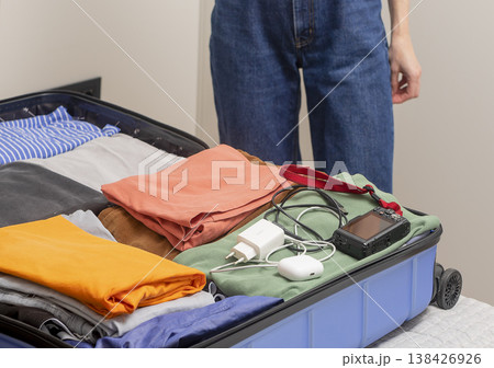 Travel gadgets and cables rest beside folded clothes during packing. Female hands place camera 138426926