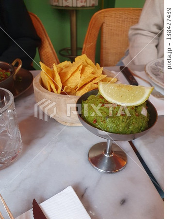 Fresh green guacamole topped with a lemon wedge served alongside crisp tortilla chips on a marble table in a cozy restaurant setting, evoking casual dining, sharing and social snacking. Fresh green guacamole topped with a lemon wedge served alongside crisp tortilla chips on a marble table in a cozy restaurant setting, evoking casual dining, sharing and social snacking. 138427439