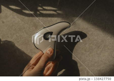 Hand holding modern microcurrent facial massager in warm sunlight with deep shadows, aesthetic beauty tech, minimalist skin care routine at home, sculptural guasha device, moody wellness concept. 138427750