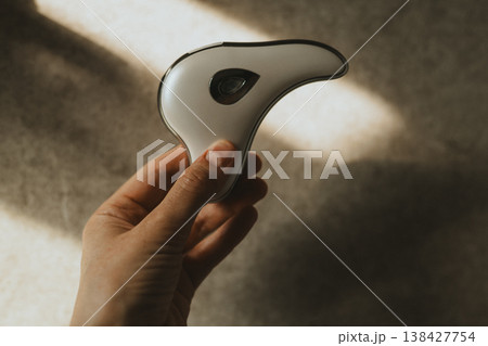 Hand holding modern microcurrent facial massager in warm sunlight with deep shadows, aesthetic beauty tech, minimalist skin care routine at home, sculptural guasha device, moody wellness concept. Hand holding modern microcurrent facial massager in warm sunlight with deep shadows, aesthetic beauty tech, minimalist skin care routine at home, sculptural guasha device, moody wellness concept. 138427754