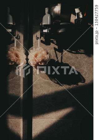 Aesthetic bathroom interior with pink mesh sponge hanging on a hook, dramatic sunlight and deep shadows on textured wall, minimalist spa concept, mindful self care and wellness atmosphere. 138427759