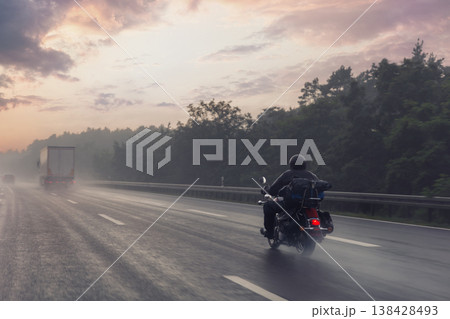 Motorcycle rider speeds along misty highway sunrise red tail light glow rain soaked asphalt drifting spray. Forest silhouette frames horizon truck fades fog evoking freedom adventure, risk solitude 138428493