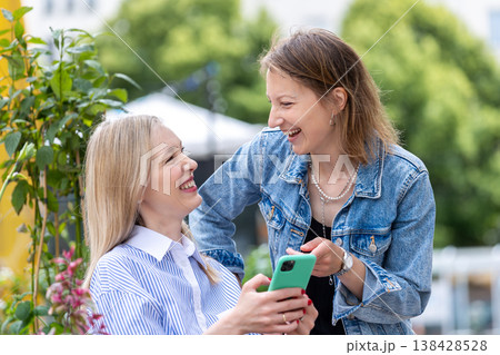Two women laugh together on sunny terrace greenery plants flowers sharing smartphone lively conversation. Casual outfits warm daylight joyful expressions friendship gossip relaxed urban lifestyle Two women laugh together on sunny terrace greenery plants flowers sharing smartphone lively conversation. Casual outfits warm daylight joyful expressions friendship gossip relaxed urban lifestyle 138428528