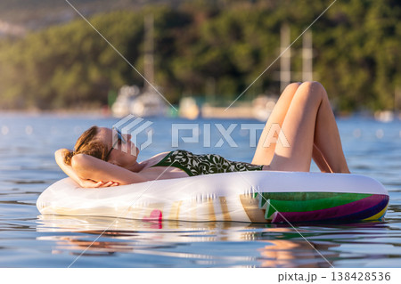 Woman wear green swimsuit relax inflatable ring calm sea near yachts forested coast sunset warm summer sunlight. Female person peaceful moment  leisure freedom vacation lifestyle serene getaway mood 138428536