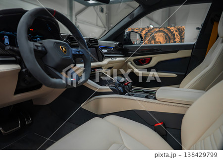 Close up Lamborghini Urus SE interior shows flat bottom wheel with bull emblem, angular vents, jet fighter toggles, cream leather seats, carbon trim, red inserts, workshop visible, soft even lighting. 138429799
