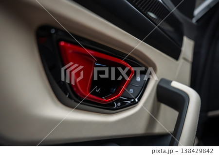 A close up shows a red triangular door handle in a glossy black bezel, with lit lock and window icons, tan leather, and a grab handle, in a Lamborghini Urus workshop. 138429842