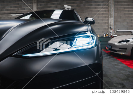 Close up of left Lamborghini Urus SE LED headlight in a premium workshop. A silver Tesla Model 3 sits on a red mat with detailing gear and checkered floor. 138429846