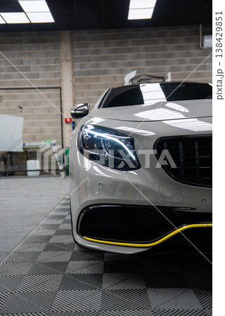 Close view shows Panamericana grille, LED headlight, and yellow bumper line on a Mercedes AMG in an auto workshop with brick walls, fluorescent panels, and checkered floor. Close view shows Panamericana grille, LED headlight, and yellow bumper line on a Mercedes AMG in an auto workshop with brick walls, fluorescent panels, and checkered floor. 138429851