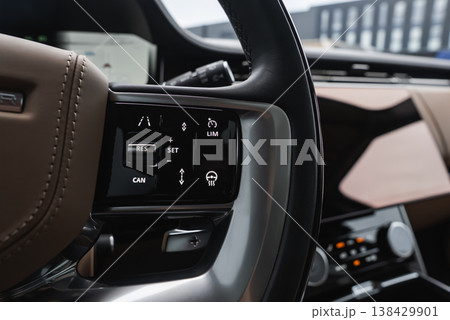 Close up interior shows brown leather wheel with RES SET, CAN, LIM, and lane keep icons. Metal rotary knobs and wood trim sit in a well lit service bay or showroom. 138429901