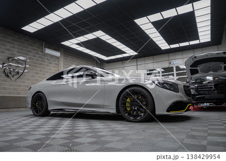 A white Mercedes AMG coupe sits on black wheels with yellow calipers in a high end workshop, checker flooring and bright panels, with another car hood open nearby. A white Mercedes AMG coupe sits on black wheels with yellow calipers in a high end workshop, checker flooring and bright panels, with another car hood open nearby. 138429954
