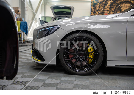 Close up of a Mercedes Benz AMG front quarter in a workshop, glossy black wheel, drilled rotors, yellow calipers, silver body with yellow trim, bright indoor lighting Close up of a Mercedes Benz AMG front quarter in a workshop, glossy black wheel, drilled rotors, yellow calipers, silver body with yellow trim, bright indoor lighting 138429997
