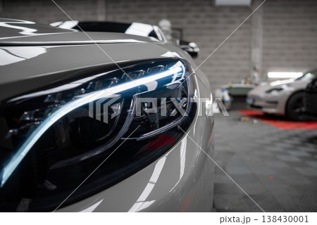Close up shows LED headlight and glossy fender of a Mercedes Benz AMG in a high end workshop, with Tesla on checkered flooring and red mats. Close up shows LED headlight and glossy fender of a Mercedes Benz AMG in a high end workshop, with Tesla on checkered flooring and red mats. 138430001