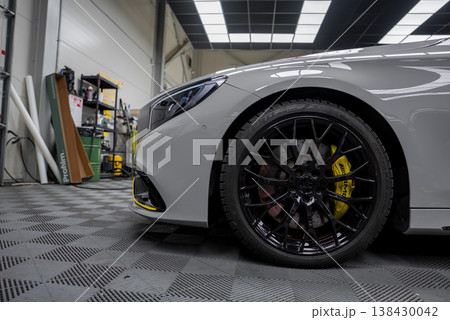 Close up low angle shows silver Mercedes Benz AMG coupe with black multi spoke wheel, yellow AMG brake caliper, and LED headlamp in an indoor detailing bay. Close up low angle shows silver Mercedes Benz AMG coupe with black multi spoke wheel, yellow AMG brake caliper, and LED headlamp in an indoor detailing bay. 138430042