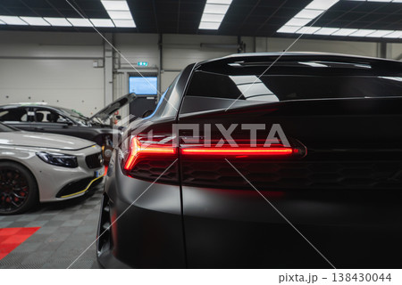 A matte black Lamborghini Urus SE shows a rear three quarter view, LED taillight lit. A white Mercedes Benz AMG coupe sits by an open hood in a bright service workshop. 138430044