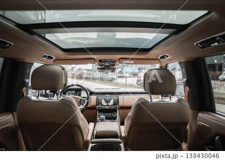 Wide angle view from second row shows tan leather seats, dual screens, and panoramic twin glass panels. Workshop or dealership lot with luxury cars is visible ahead. 138430046