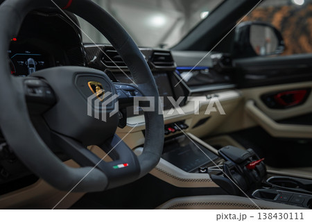 Stationary Lamborghini Urus SE close up shows suede steering wheel with bull emblem, tricolor badge, digital cluster, angular vents, touchscreen, and jet style selector. 138430111