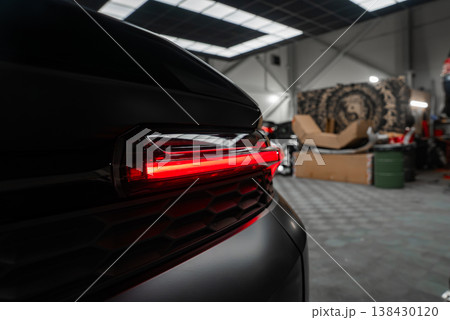 A close up shows the left rear LED taillight glowing on a black luxury SUV inside a high end workshop with skylights, tools, large wheels, and boxes in low key light. 138430120