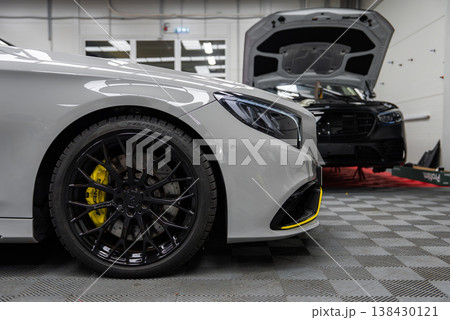 Close up shows a white Mercedes Benz AMG coupe with LED headlamp, black multi spoke wheel, and yellow accents. A black Range Rover on a lift sits behind under bright lights. 138430121