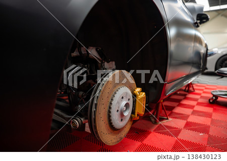 Close up of a luxury SUV on jack stands with front wheel off, showing drilled rotor and gold caliper on a red checkered floor in a professional service bay, low angle. Close up of a luxury SUV on jack stands with front wheel off, showing drilled rotor and gold caliper on a red checkered floor in a professional service bay, low angle. 138430123