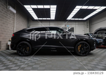 A matte black Lamborghini Urus SE sits in a modern workshop with checkered floor, grid lights, Y spoke wheels, and gold calipers as luxury cars undergo service nearby. 138430129